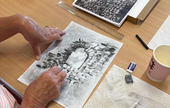A close-up view of someone drawing a stone ruin with an English Heritage branded coffee cup on the table. 