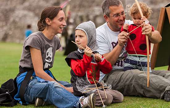 Two adults sit on the grass while two children dressed as knights play with toy bows and arrows