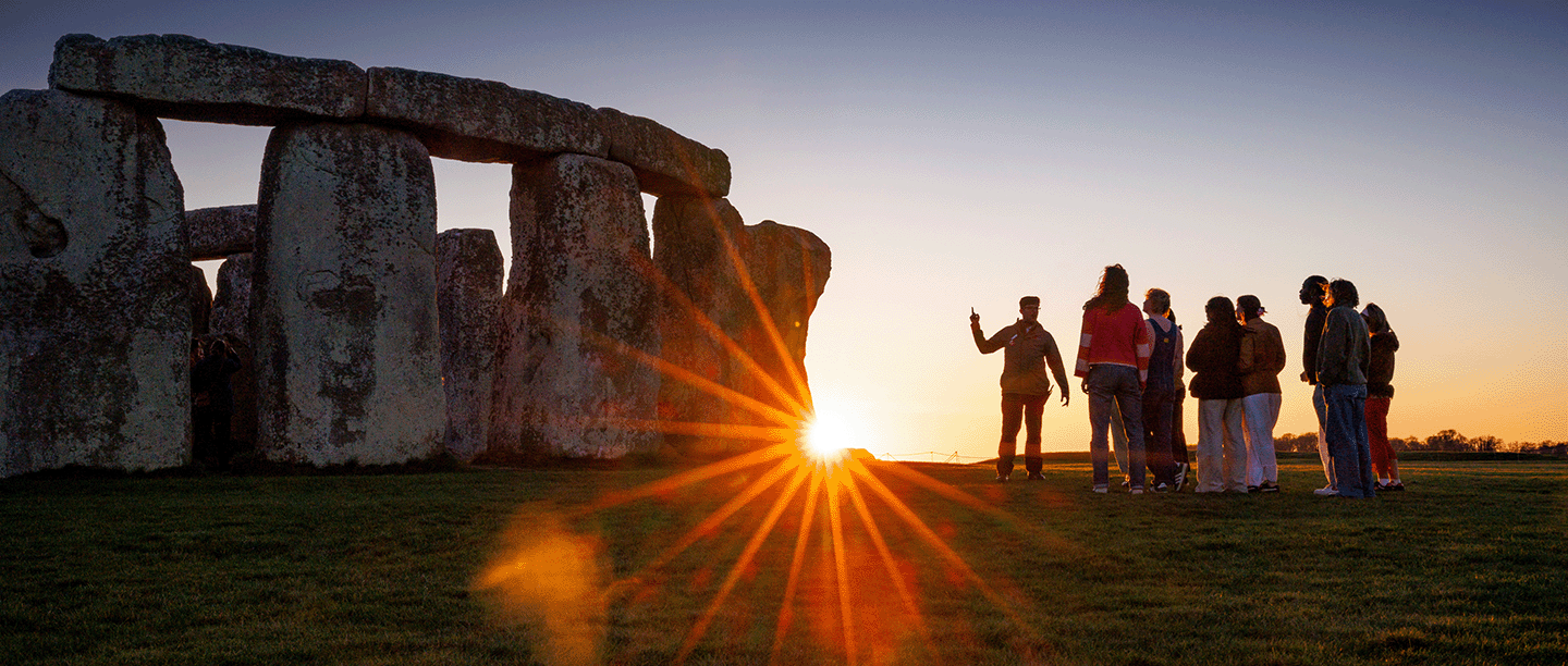 Photo of an English Heritage guide speaking to a group of visitors at Stonehenge with the sun shining through just above the ground